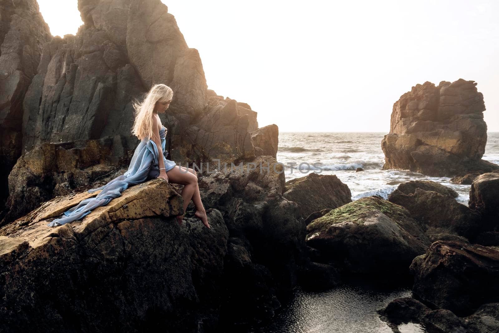 Beautiful European girl in a long dress sitting on the edge of a cliff and looking at the sea