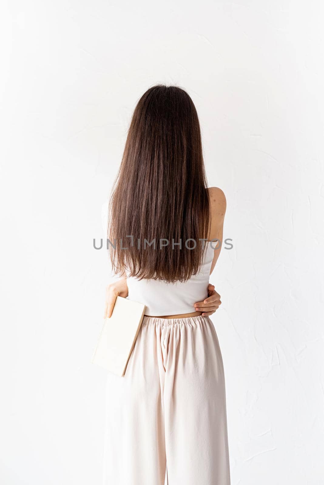 beautiful young woman with long hair in white cozy clothes on white background holding a book, view from behind by Desperada