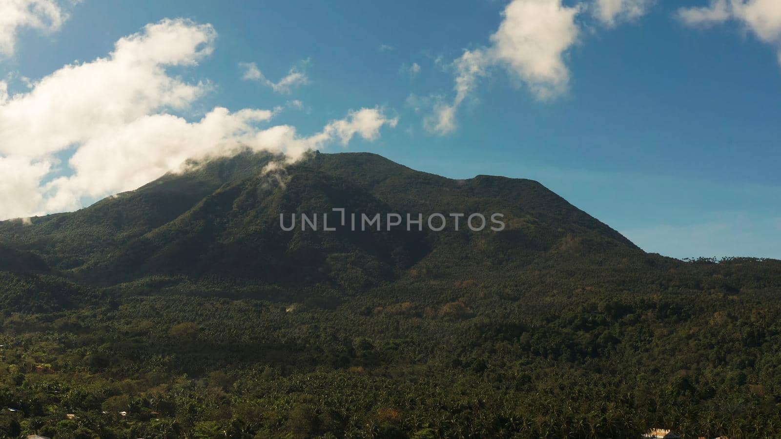 Mountains covered rainforest, trees and blue sky with clouds, aerial view. Camiguin, Philippines. Mountain landscape on tropical island with mountain peaks covered with forest. Slopes of mountains with evergreen vegetation.