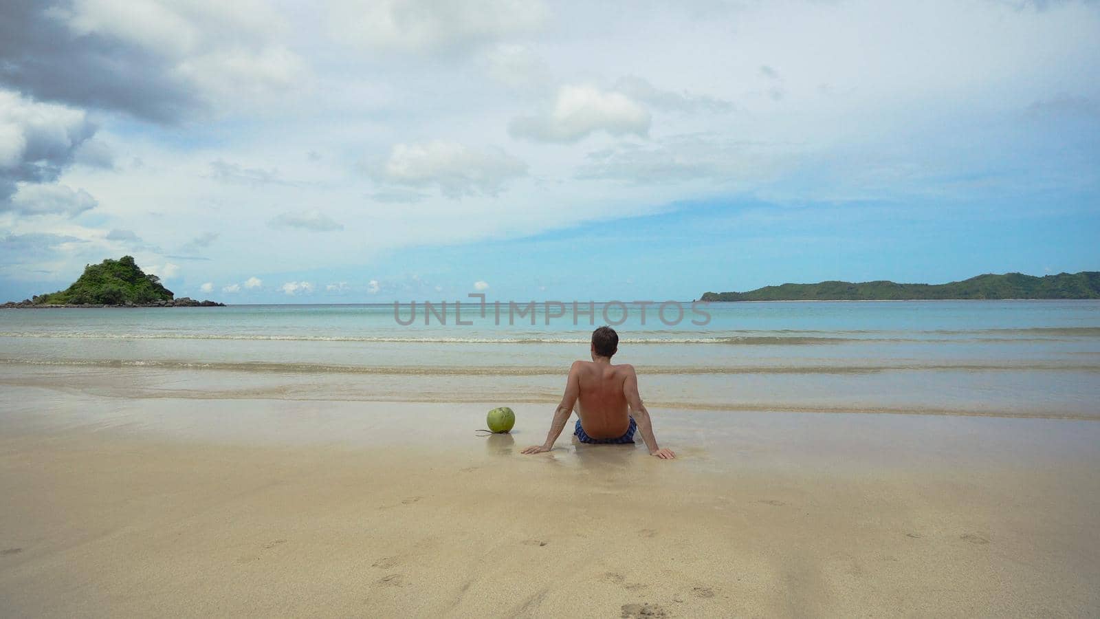 Man sits on a sandy beach and enjoy the seascape. Man relaxing sitting down on the shore of a white sand beach, with the waves bathing the shore in an idyllic nature location, being thoughtful.