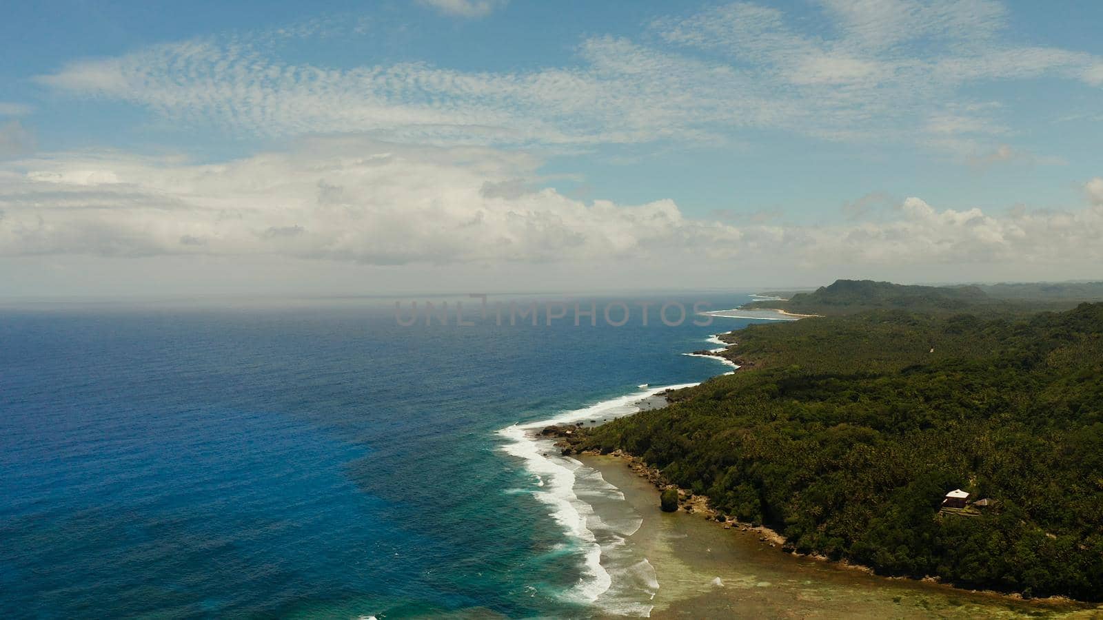 The coast of Siargao island with beach, green forest and ocean surf with waves against the sky and clouds, aerial view. tropical landscape.