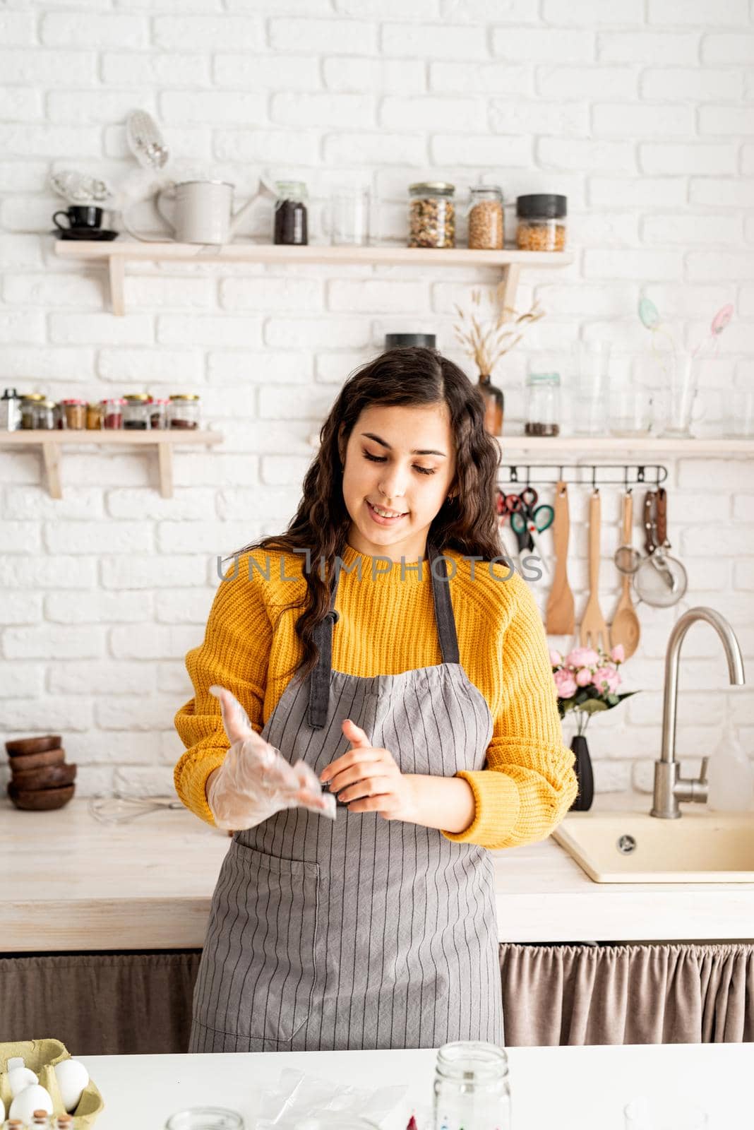 Beautiful brunette woman in yellow sweater and gray apron preparing to color easter eggs in the kitchen putting on the gloves