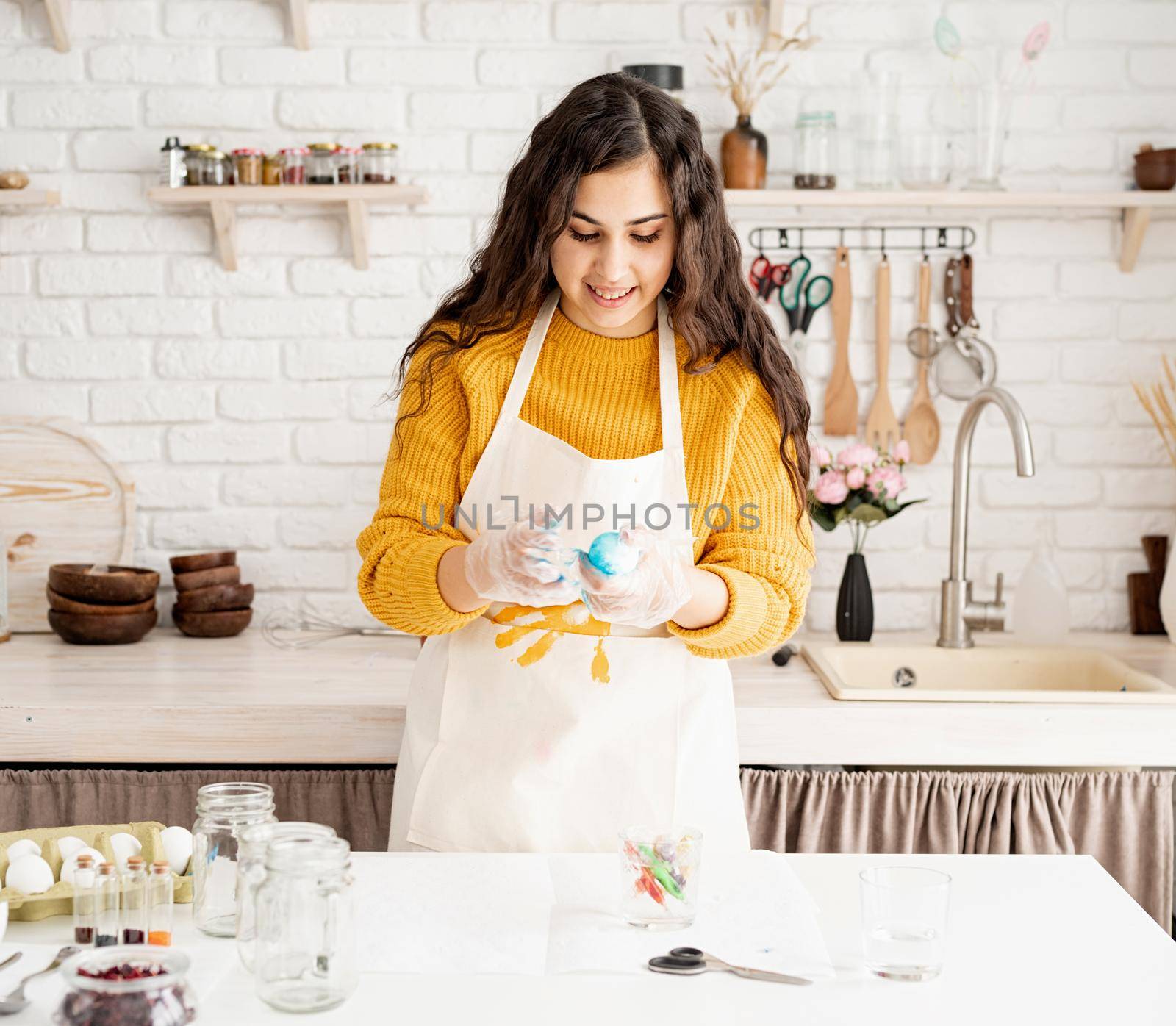 Beautiful brunette woman in yellow sweater and gray apron coloring easter eggs blue in the kitchen