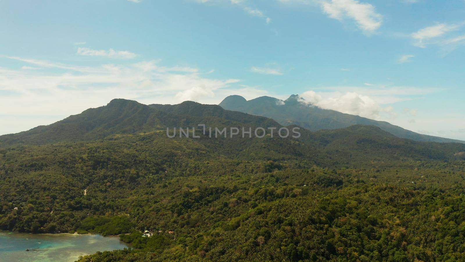 Tropical landscape: Mountains with forest, against blue sea with blue sky with clouds, top view, Camiguin, Philippines. Mountain landscape on tropical island with mountain peaks covered with forest. Slopes of mountains with evergreen vegetation.