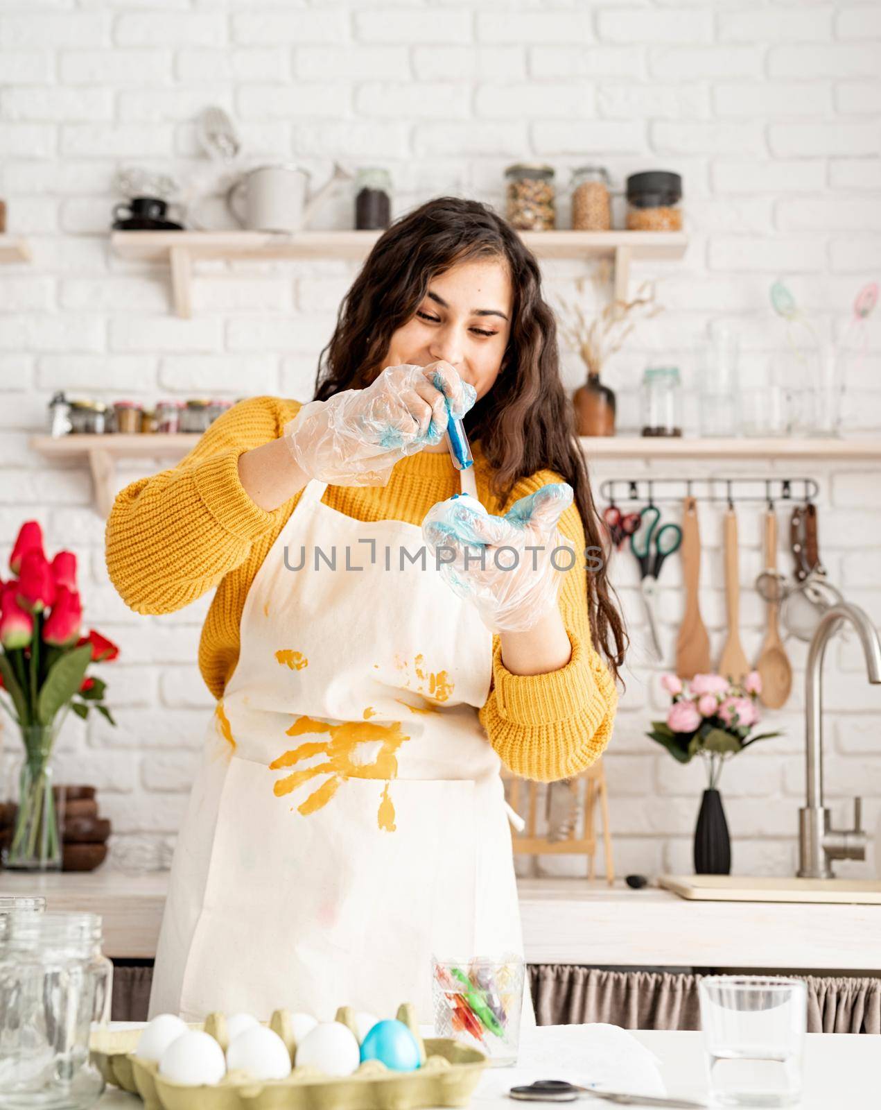 Beautiful brunette woman in yellow sweater and gray apron coloring easter eggs blue in the kitchen