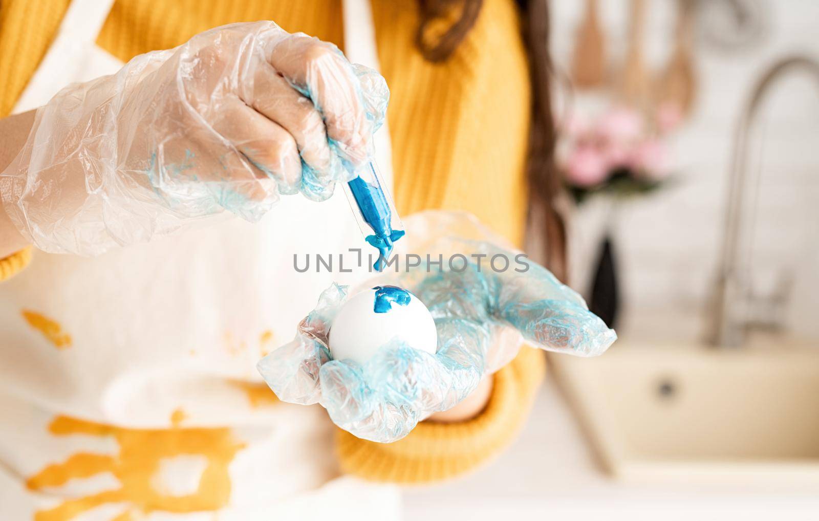 Beautiful brunette woman in yellow sweater and gray apron coloring easter eggs blue in the kitchen
