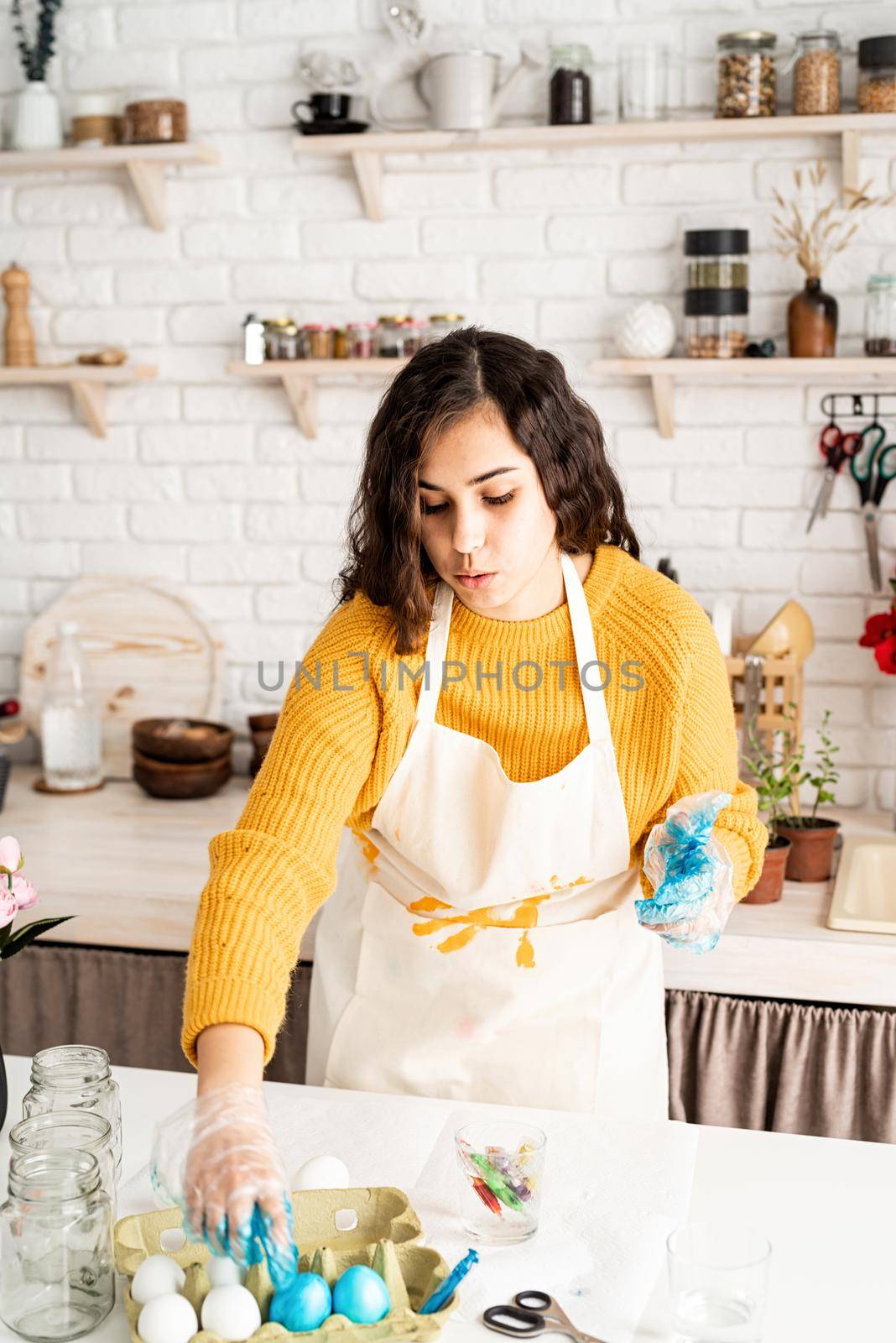 Beautiful brunette woman in yellow sweater and gray apron coloring easter eggs blue in the kitchen