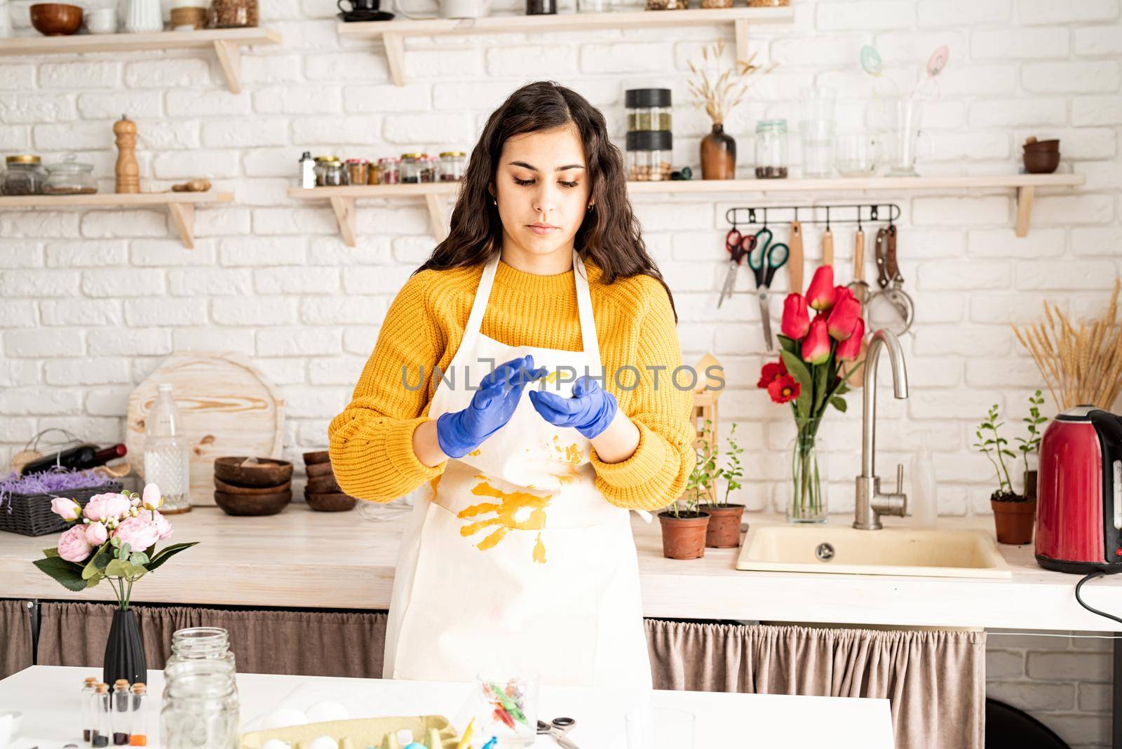 Beautiful brunette woman in yellow sweater and white apron coloring easter eggs in the kitchen