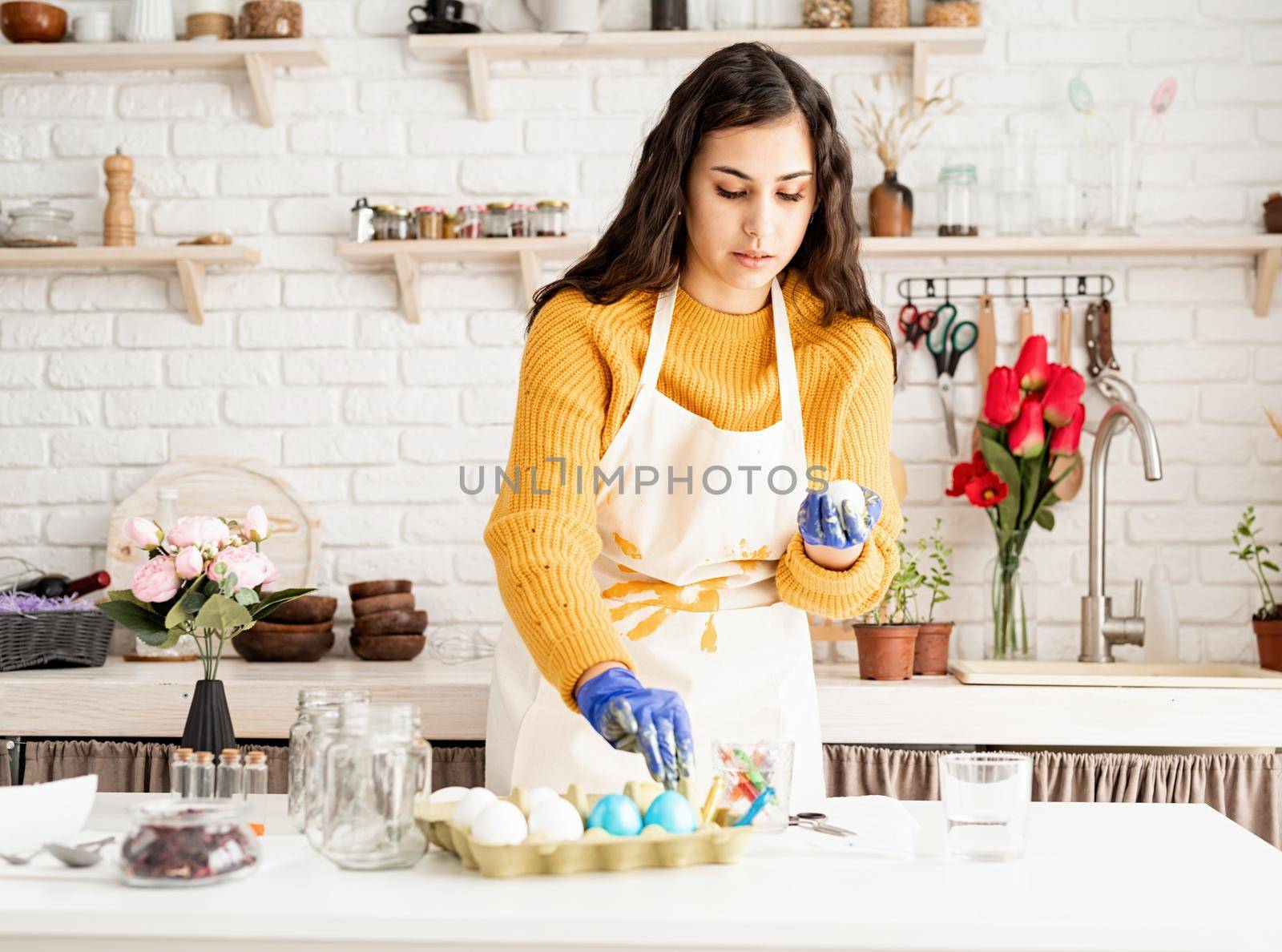 Beautiful brunette woman in yellow sweater and white apron coloring easter eggs in the kitchen
