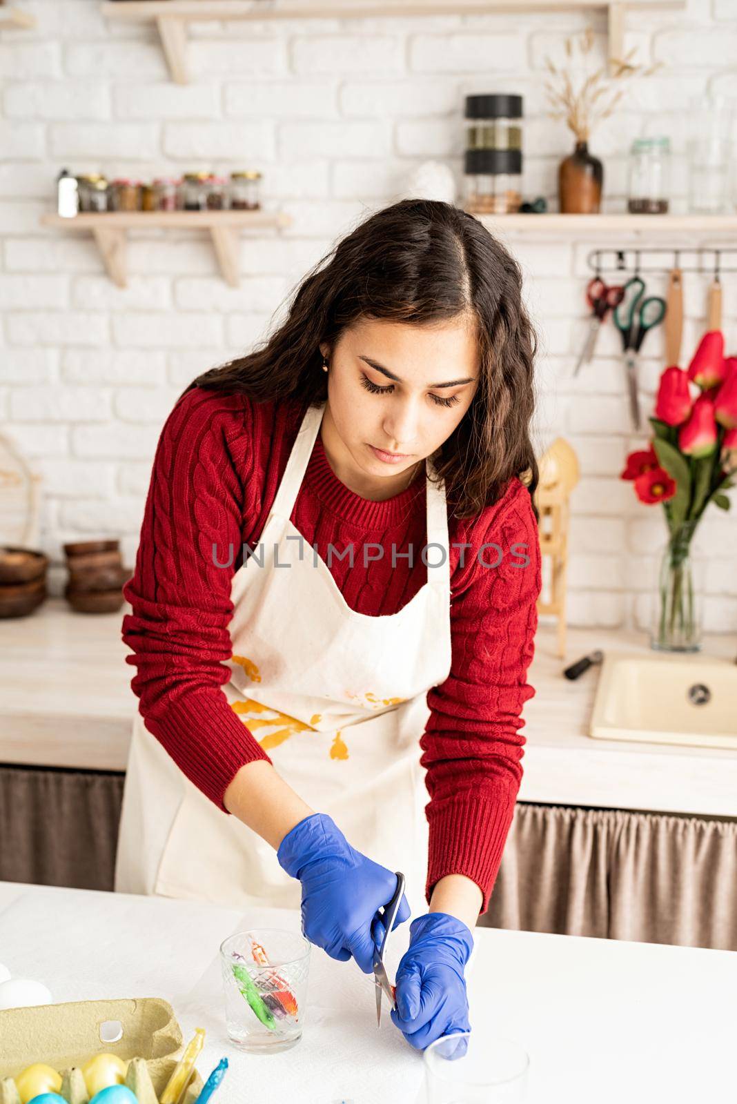 Beautiful brunette woman in red sweater and white apron coloring easter eggs in the kitchen
