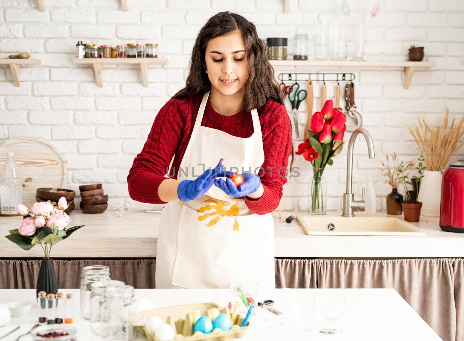 Beautiful brunette woman in red sweater and white apron coloring easter eggs in the kitchen