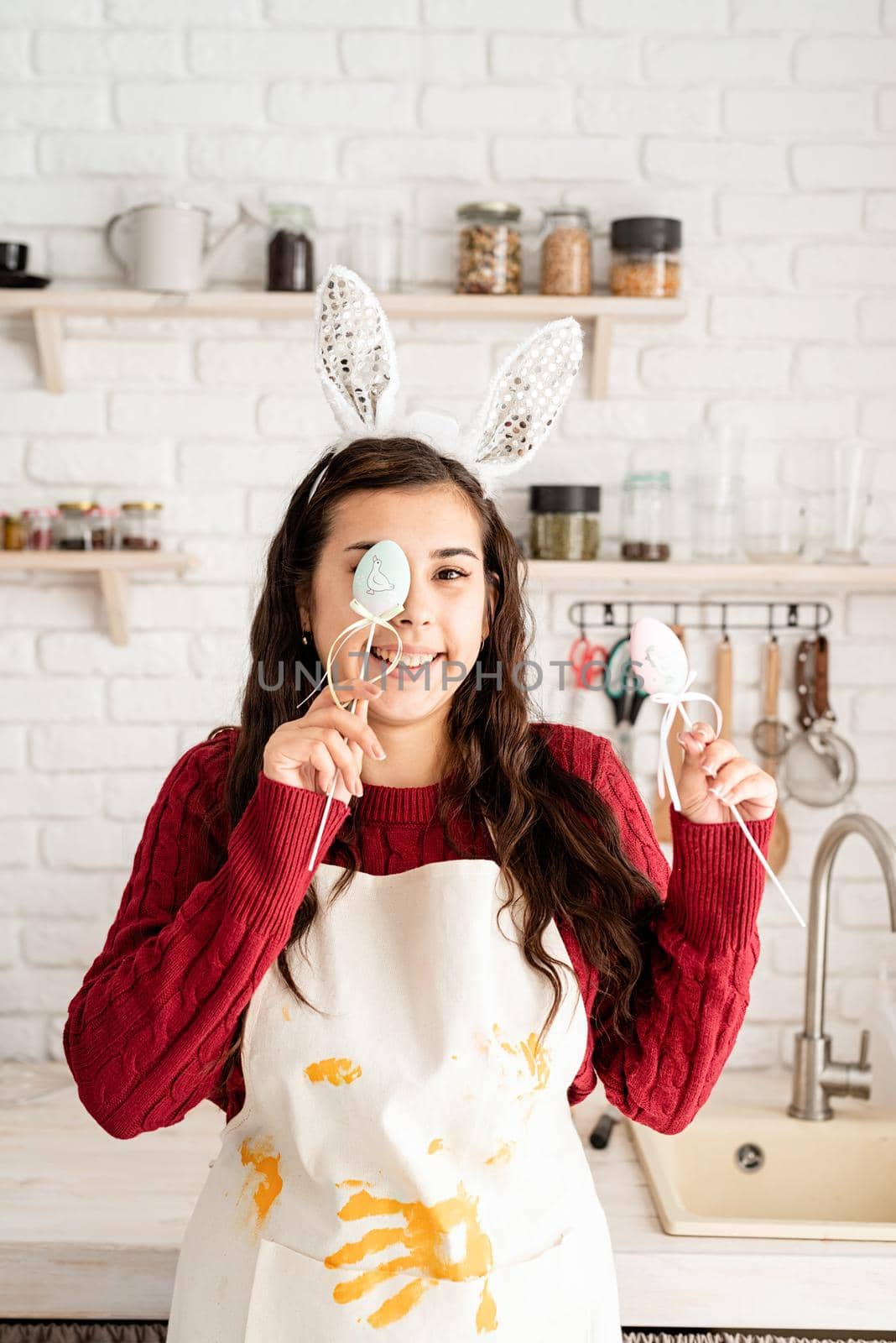 Beautiful funny brunette woman in red sweater and white apron wearing rabbit ears, covering eyes with easter eggs decorations