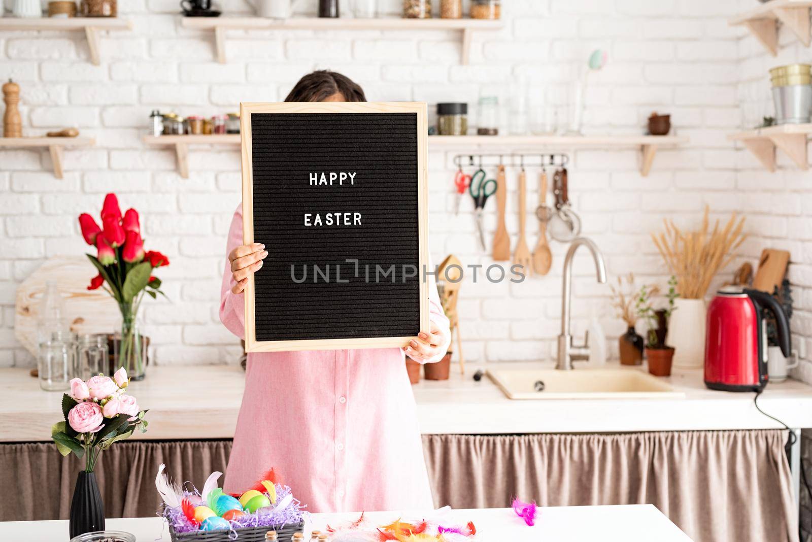 woman in pink dress holding black letter board with words Happy Easter, standing in the kitchen