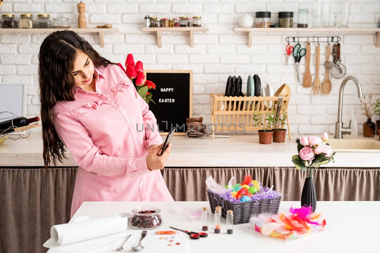 Beautiful brunette woman taking photo of decorated easter eggs using mobile phone in the kitchen