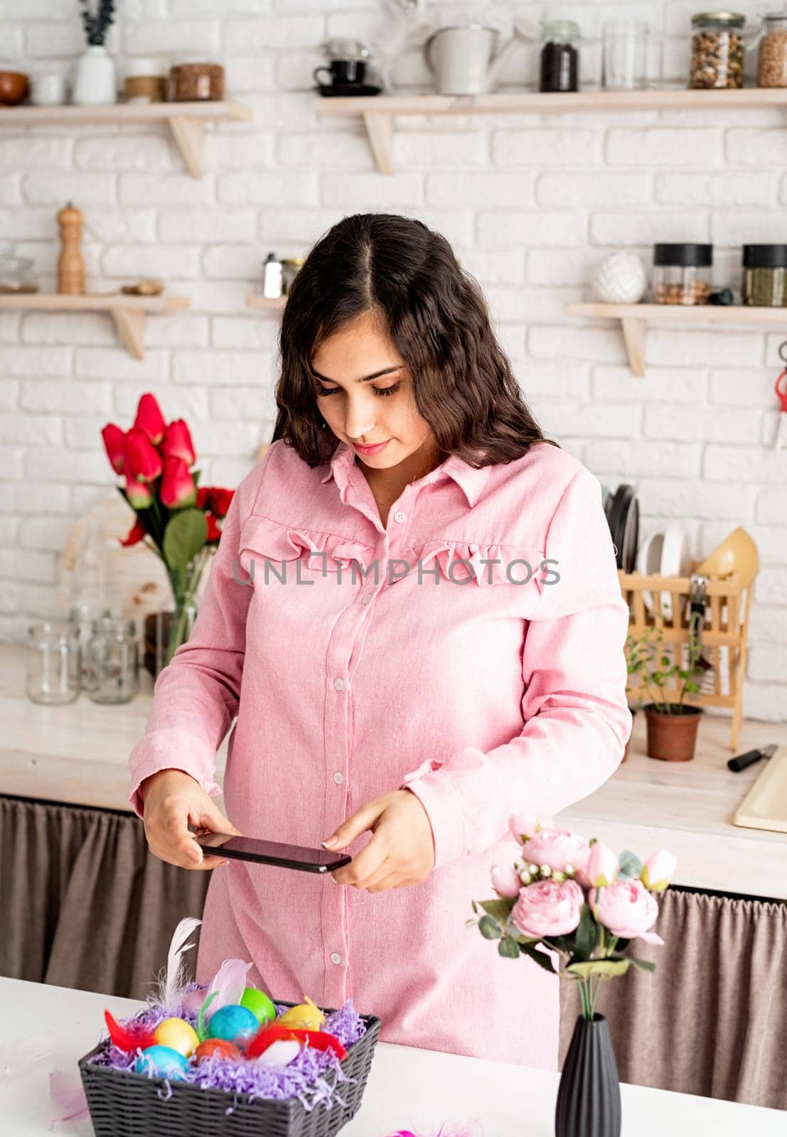 Beautiful brunette woman taking photo of decorated easter eggs using mobile phone in the kitchen