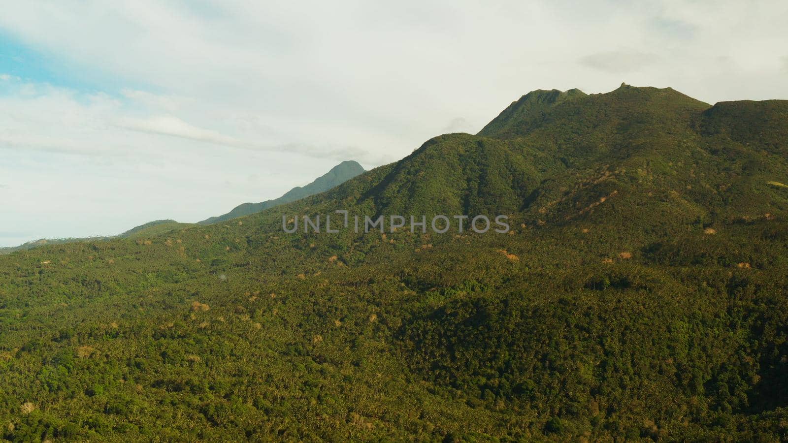 Mountains covered rainforest, trees and blue sky with clouds, aerial view. Camiguin, Philippines. Mountain landscape on tropical island with mountain peaks covered with forest. Slopes of mountains with evergreen vegetation.