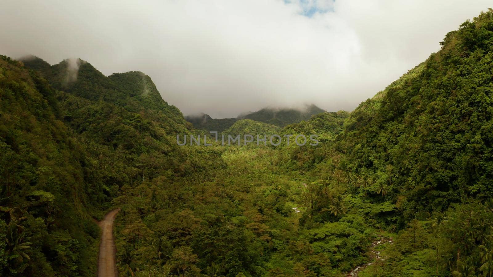 Mountains covered rainforest, trees and river in cloudy weather, aerial drone. Camiguin, Philippines. Mountain landscape on tropical island with mountain peaks covered with forest. Slopes of mountains with evergreen vegetation.