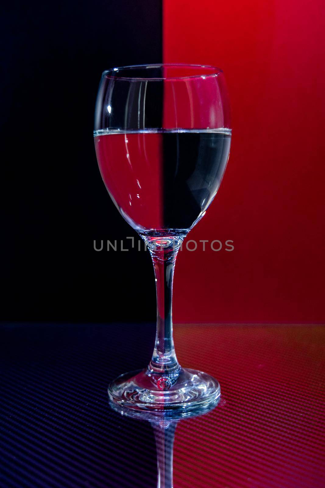 Small glassy goblet with water standing on glassy carbon reflecting surface with reflecting red and black background
