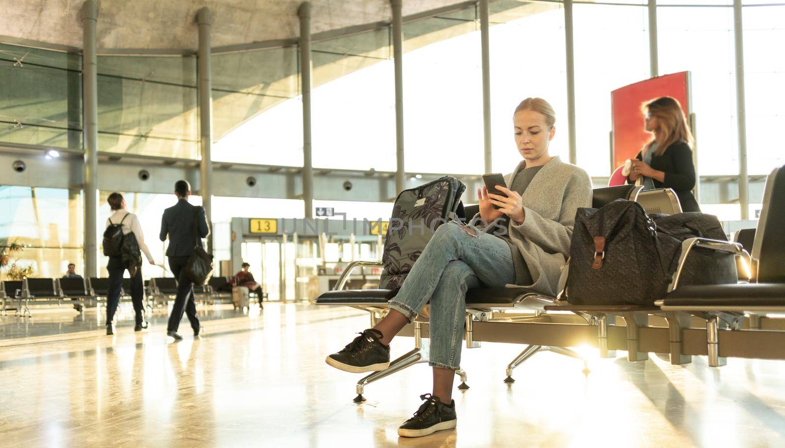 Casual blond young woman using her cell phone while waiting to board a plane at airport departure gates. by kasto