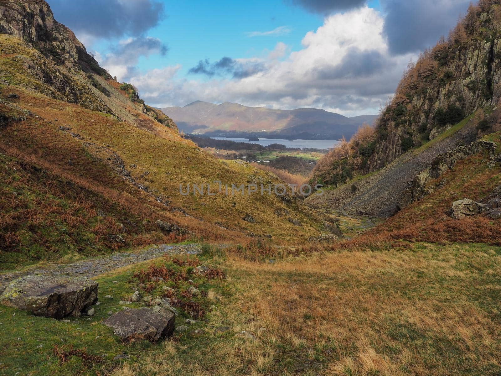 A glimpse of Derwent Water from a path winding through a steep valley with sunlight catching the fells in the distance, Lake District, UK