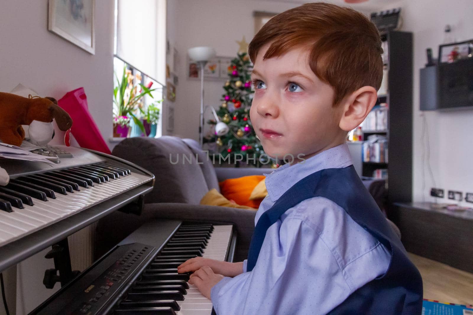 A cute, blue-eyed, red-haired, 4 years old boy playing the piano in a living room with a Christmas tree in thee background