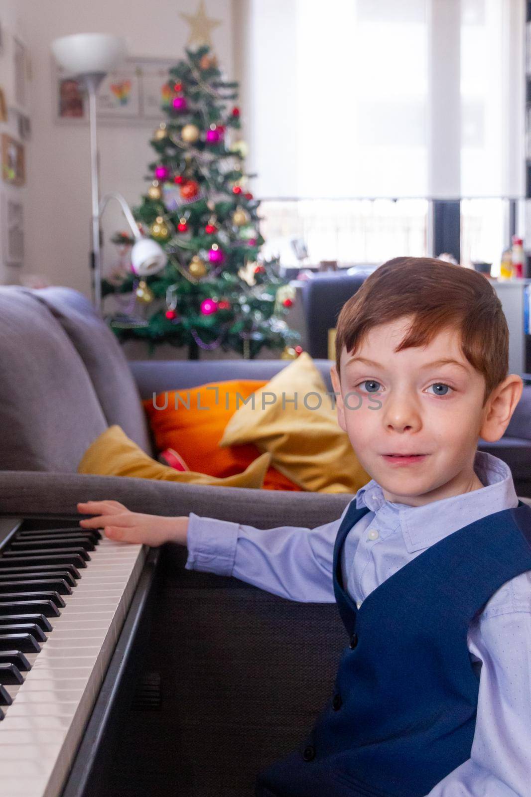 A cute, blue-eyed, red-haired, 4 years old boy playing the piano in a living room with a Christmas tree in thee background