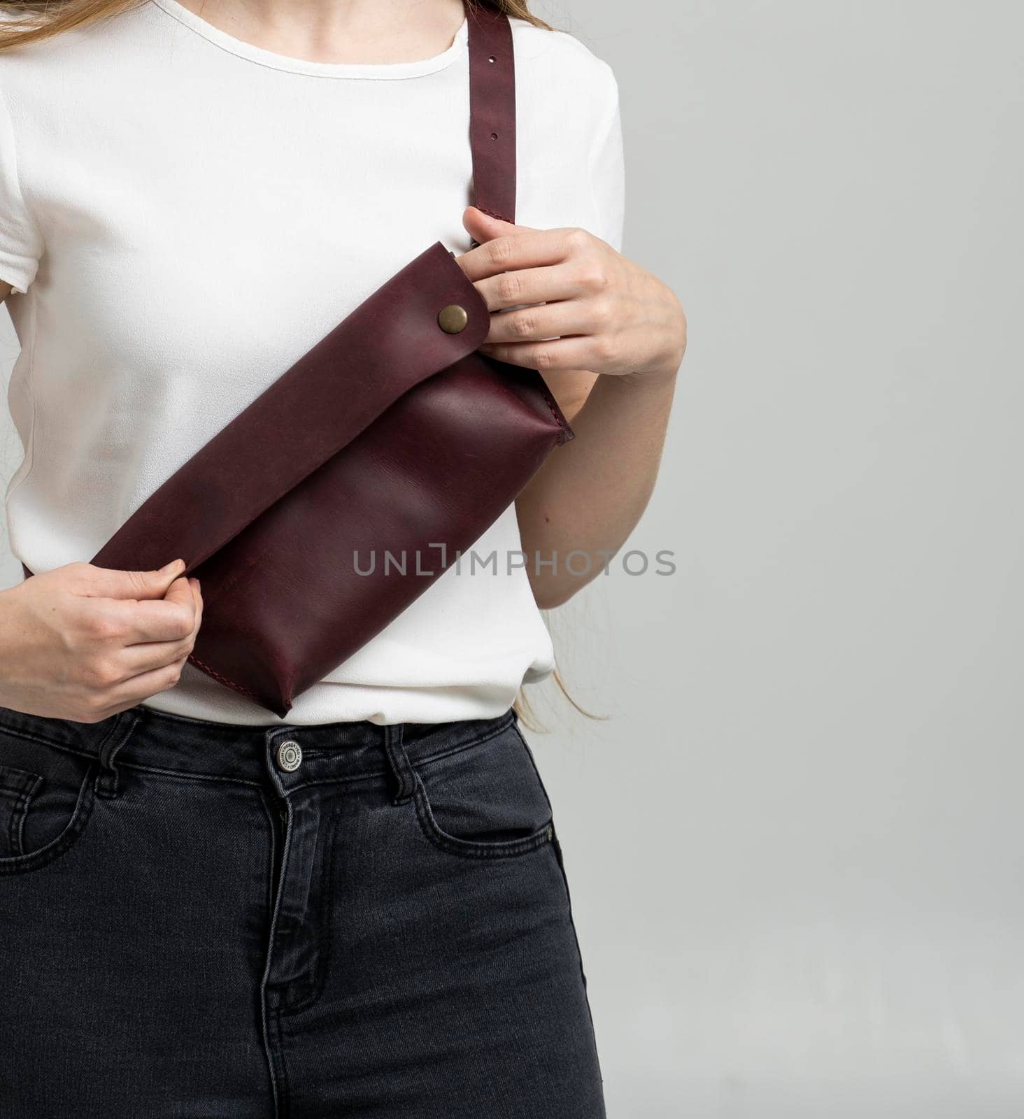 Girl in a white blouse with a leather red handmade bag over her shoulder. Designer dark red banana bag. Woman in a studio. Comfortable small bag for walking