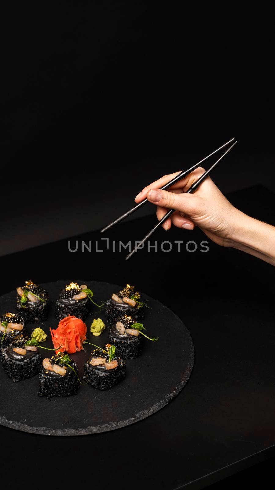 Hand with chopsticks wants to take custom sushi roll with black rice, crab meat, avocado, smoked salmon mousse, oar caviar, masago, shrimp cocktail, edible gold leaf, ginger, wasabi on black table