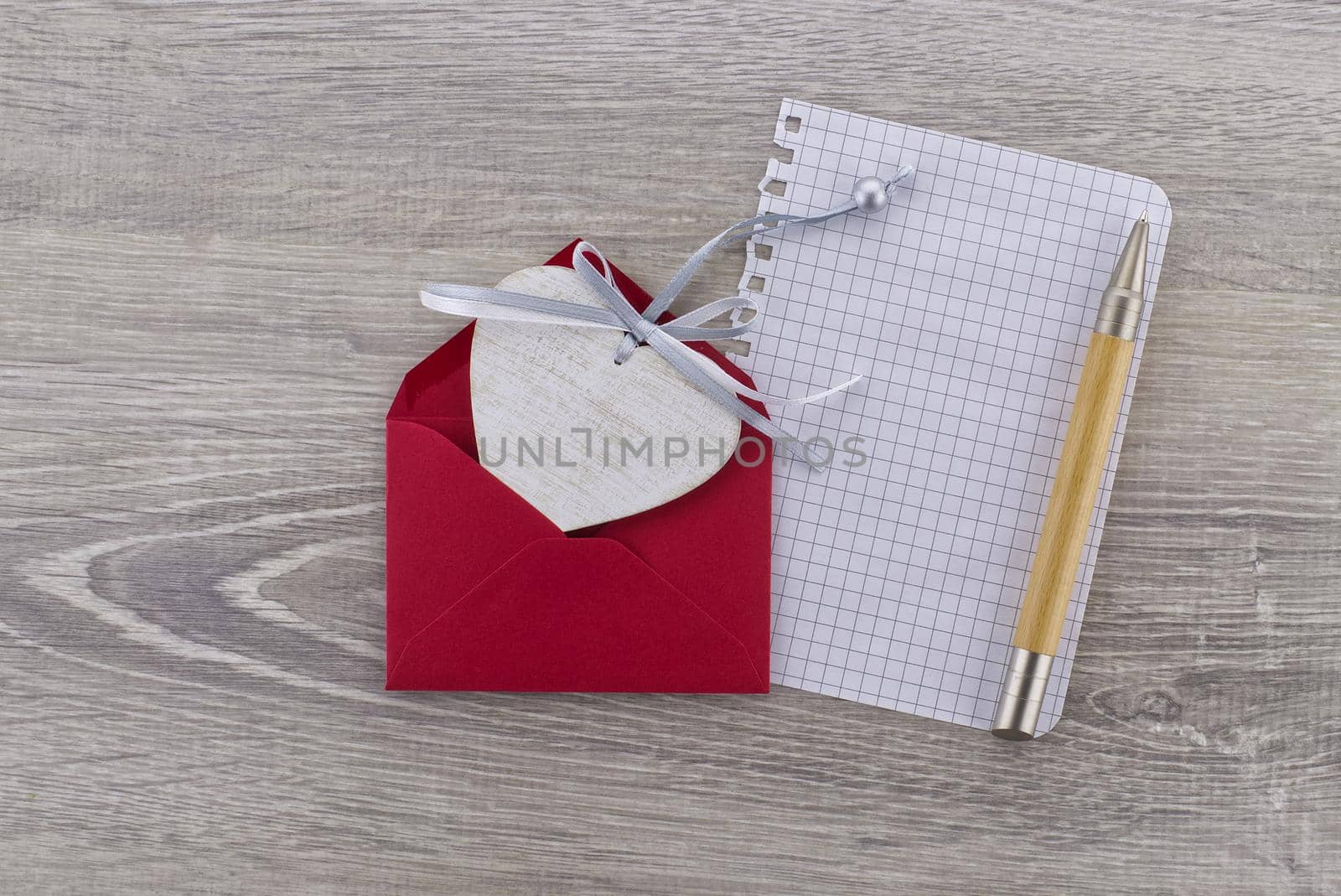 Heart-shaped wooden medallion placed in a red envelope next to a sheet of paper with a pen