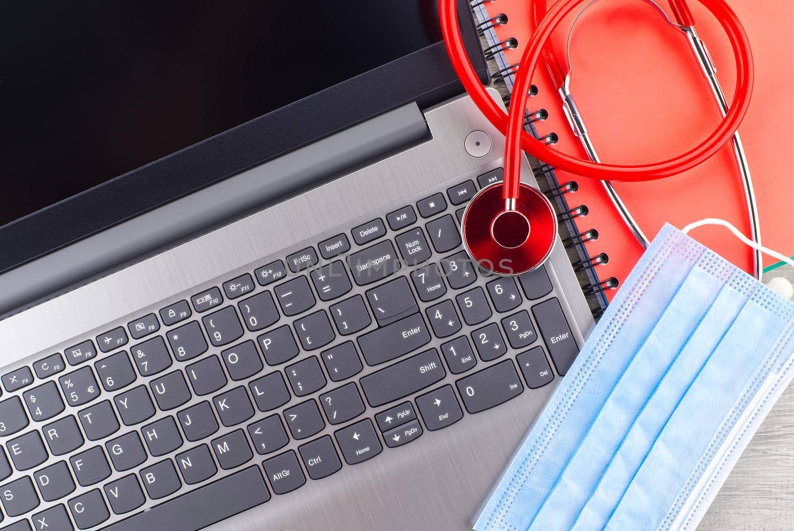 Virus pandemic and medical concept with a colorful red stethoscope coiled over a surgical face mask alongside a notebook on a wooden desk with computer keyboard viewed top down