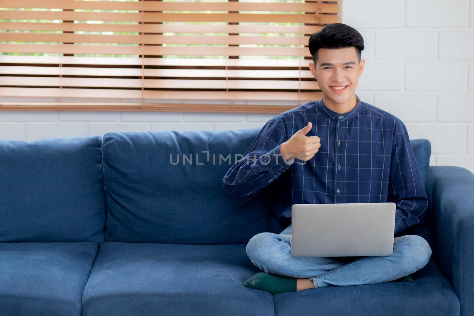 Young asian businessman smile and work from home with laptop computer online to internet on sofa in living room, freelance man using notebook on couch with comfort, new normal, lifestyle concept.