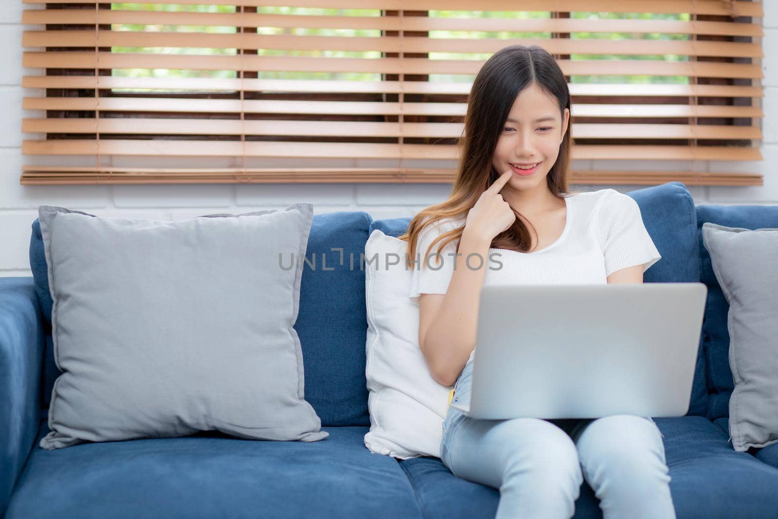 Beautiful young asian woman working on laptop computer and thinking idea on sofa at home, freelance girl sitting on couch using notebook to internet at living room, one person, lifestyle concept.