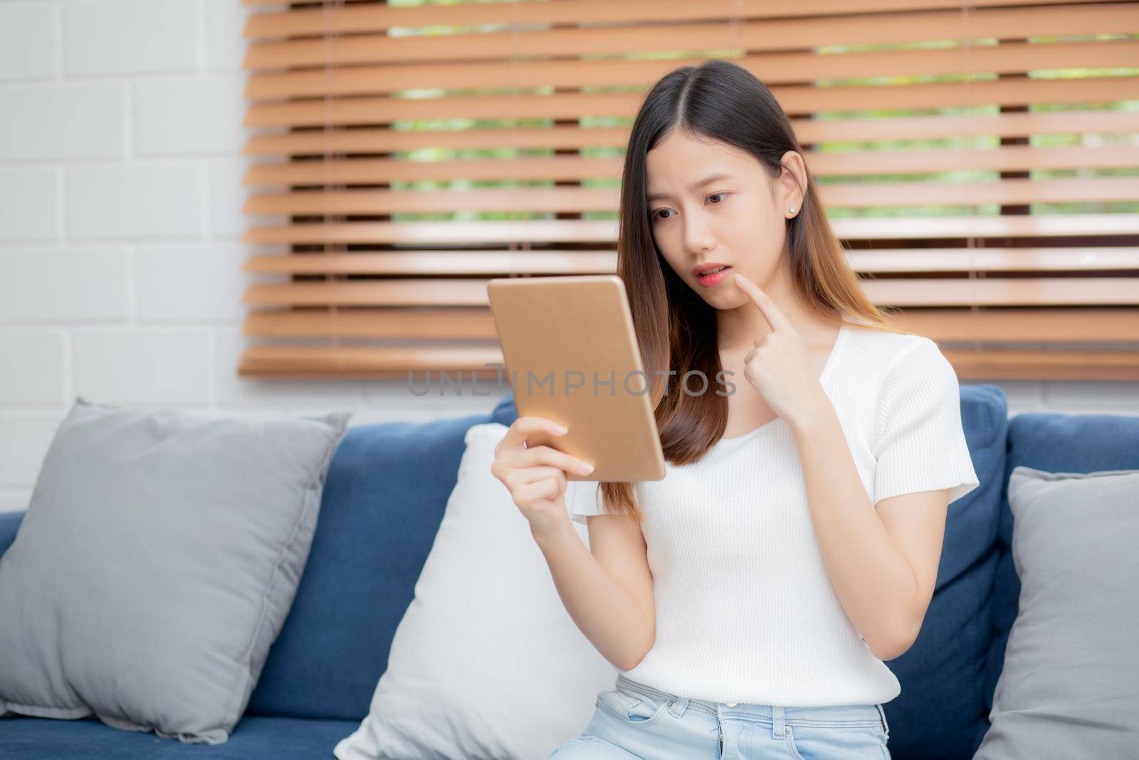 Young attractive asian woman resting using browsing tablet computer on sofa at home, happy girl sitting on couch relax looking digital gadget with excited, communication and lifestyle concept.