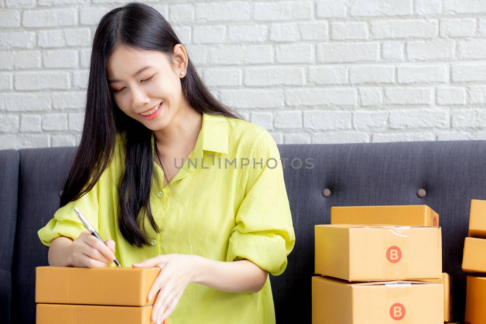 Young asian woman writing address on parcel box at home for delivery order to customer, shipping and logistic, merchant online and seller, business owner or SME, online shopping and e-commerce.