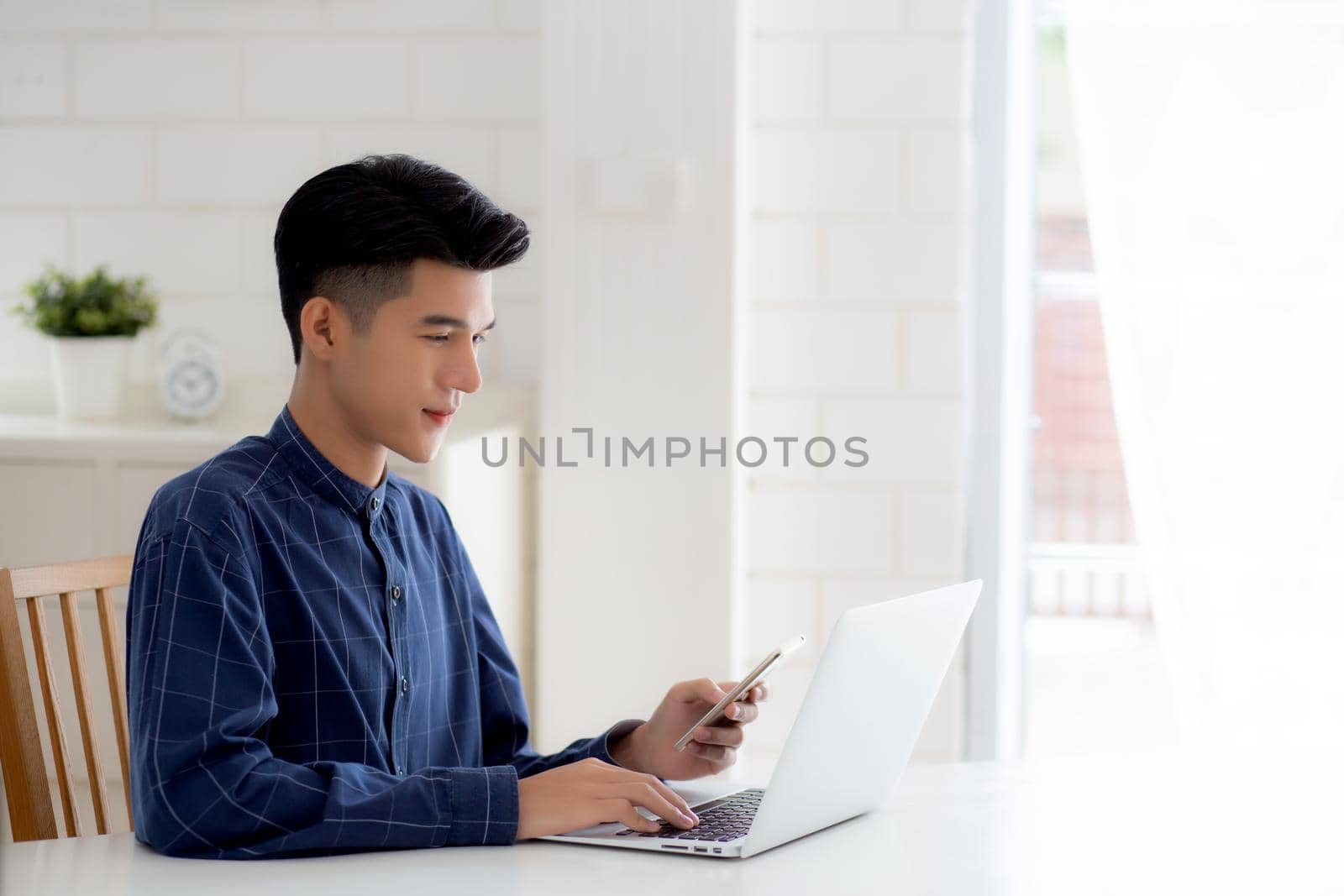 Young asian man working laptop computer and reading smartphone on internet online on desk at home, freelance male smiling using phone with social media on table, business and communication concept.