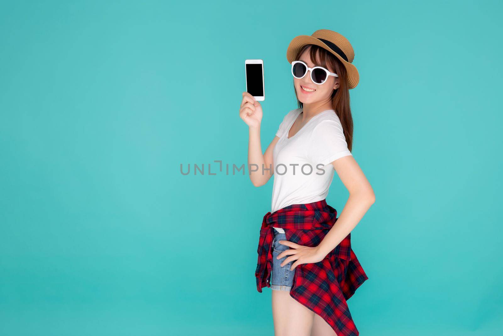 Beautiful young asian woman showing smart mobile phone for presenting something in travel summer trip in vacation isolated on blue background, asia girl holding smartphone, holiday concept.