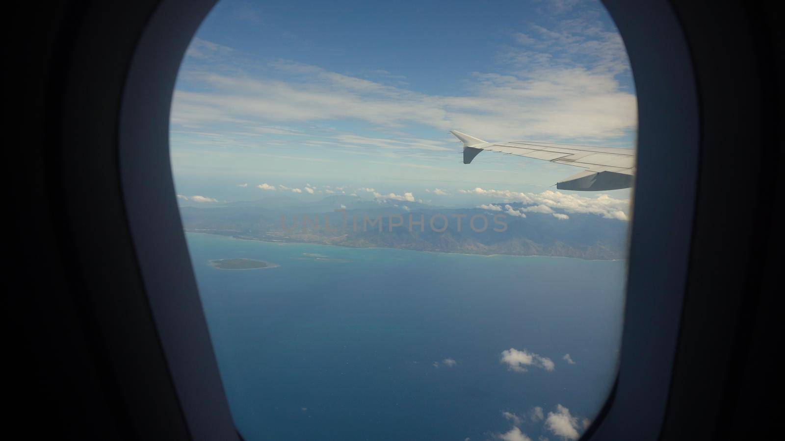 Airplane flying over blue sea and tropical island. Airplane wing through the porthole. Looking through window aircraft during flight