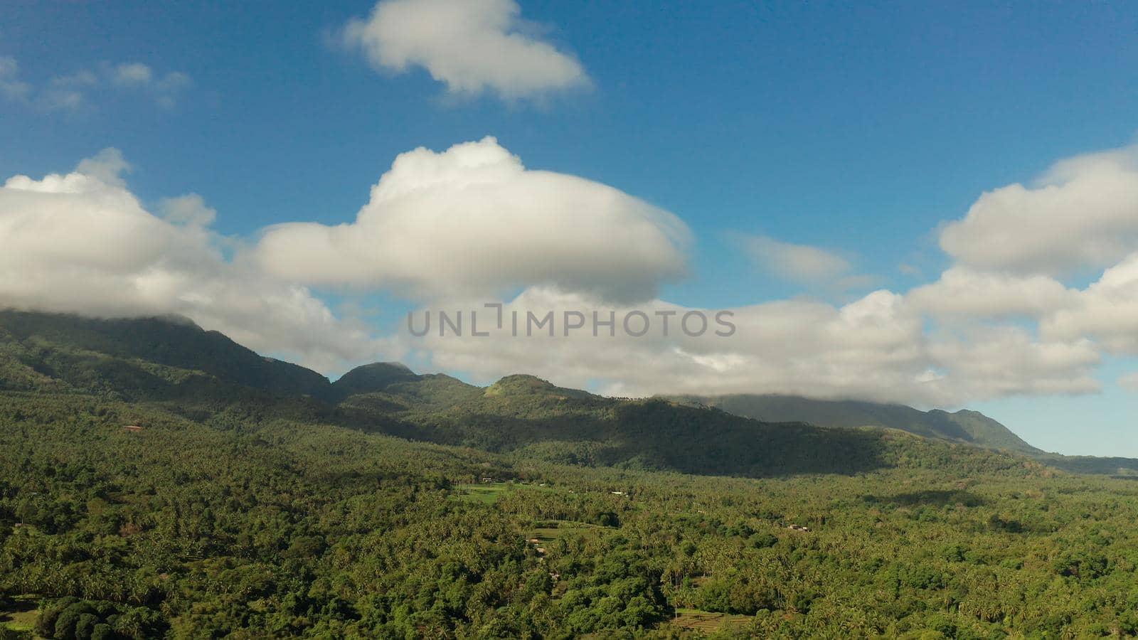 Mountains covered rainforest, trees and blue sky with clouds, aerial view. Camiguin, Philippines. Mountain landscape on tropical island with mountain peaks covered with forest. Slopes of mountains with evergreen vegetation.