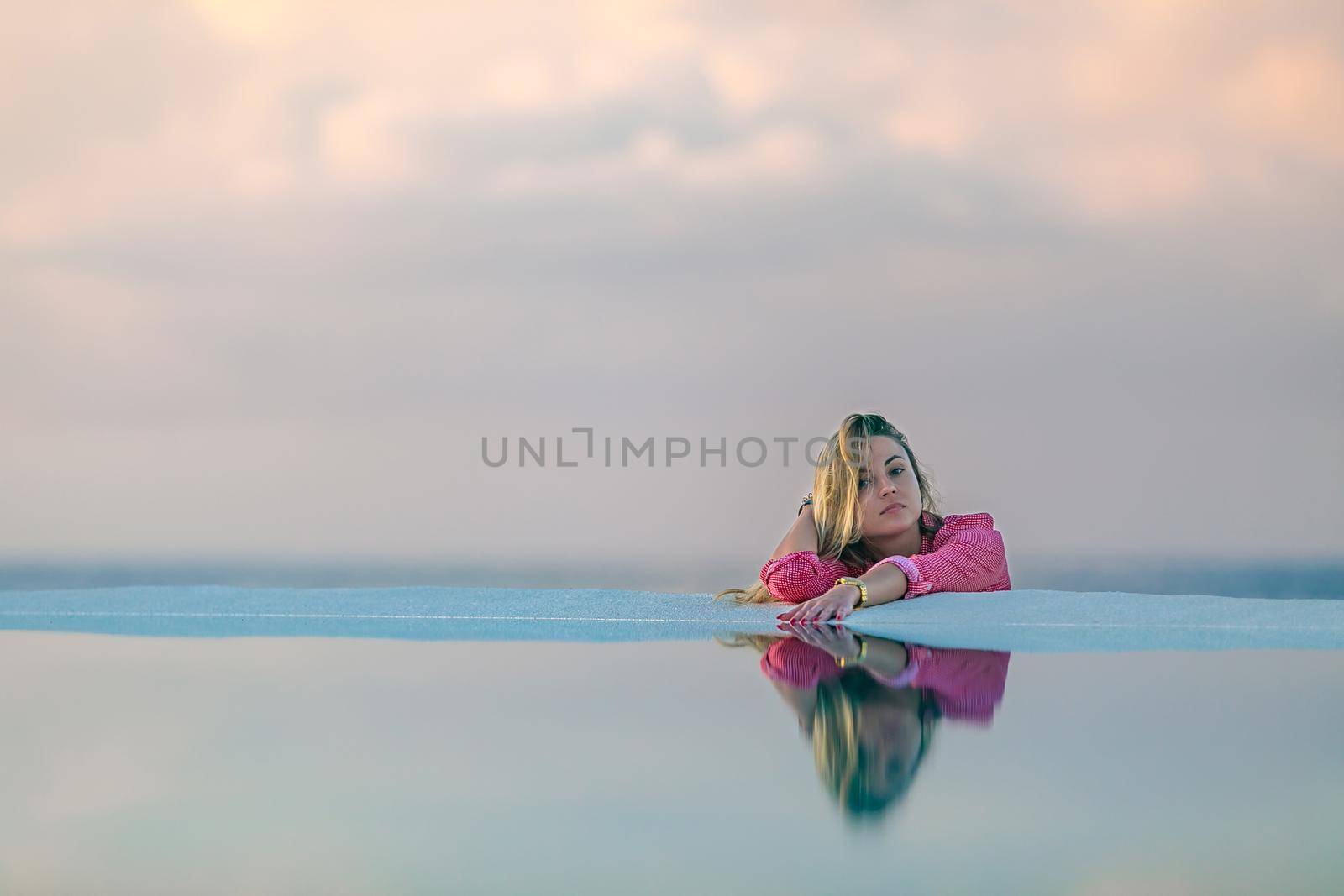 A young girl in a red shirt, at dawn by the sea pool, calm and relaxed looking at the reflection in the water. Pink clouds in the background.