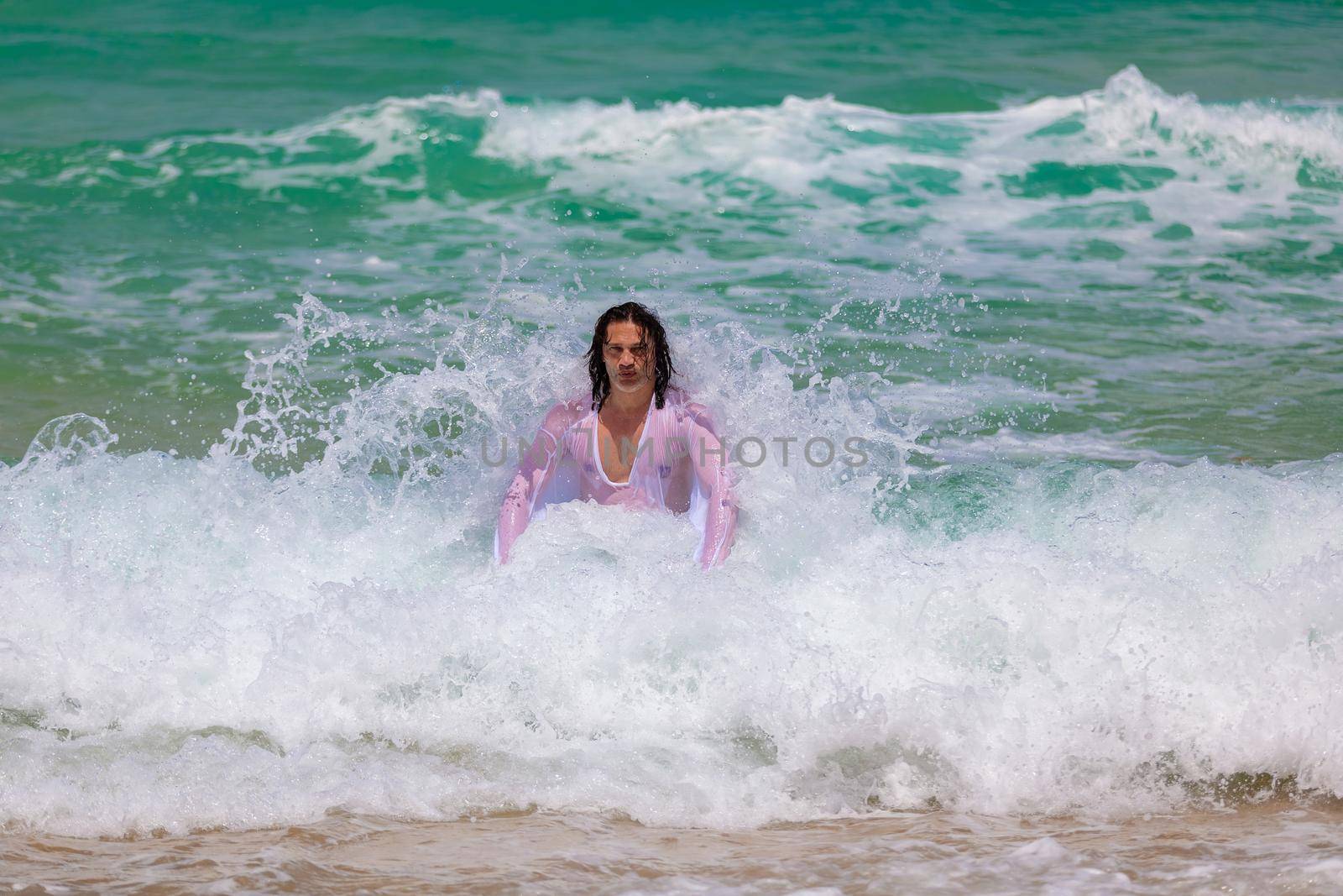 A man with long hair in a white shirt resists powerful strong and foamy waves. The spray hits him in the back
