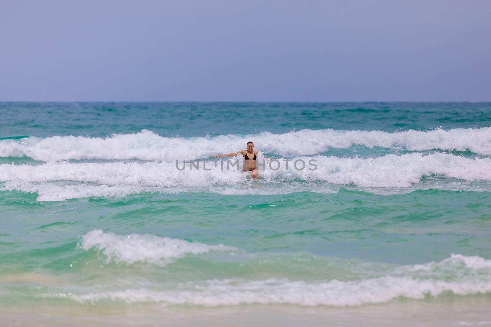 Young beautiful girl enjoys the sea and the waves, laughing raising her hands up in a black fashionable swimsuit.