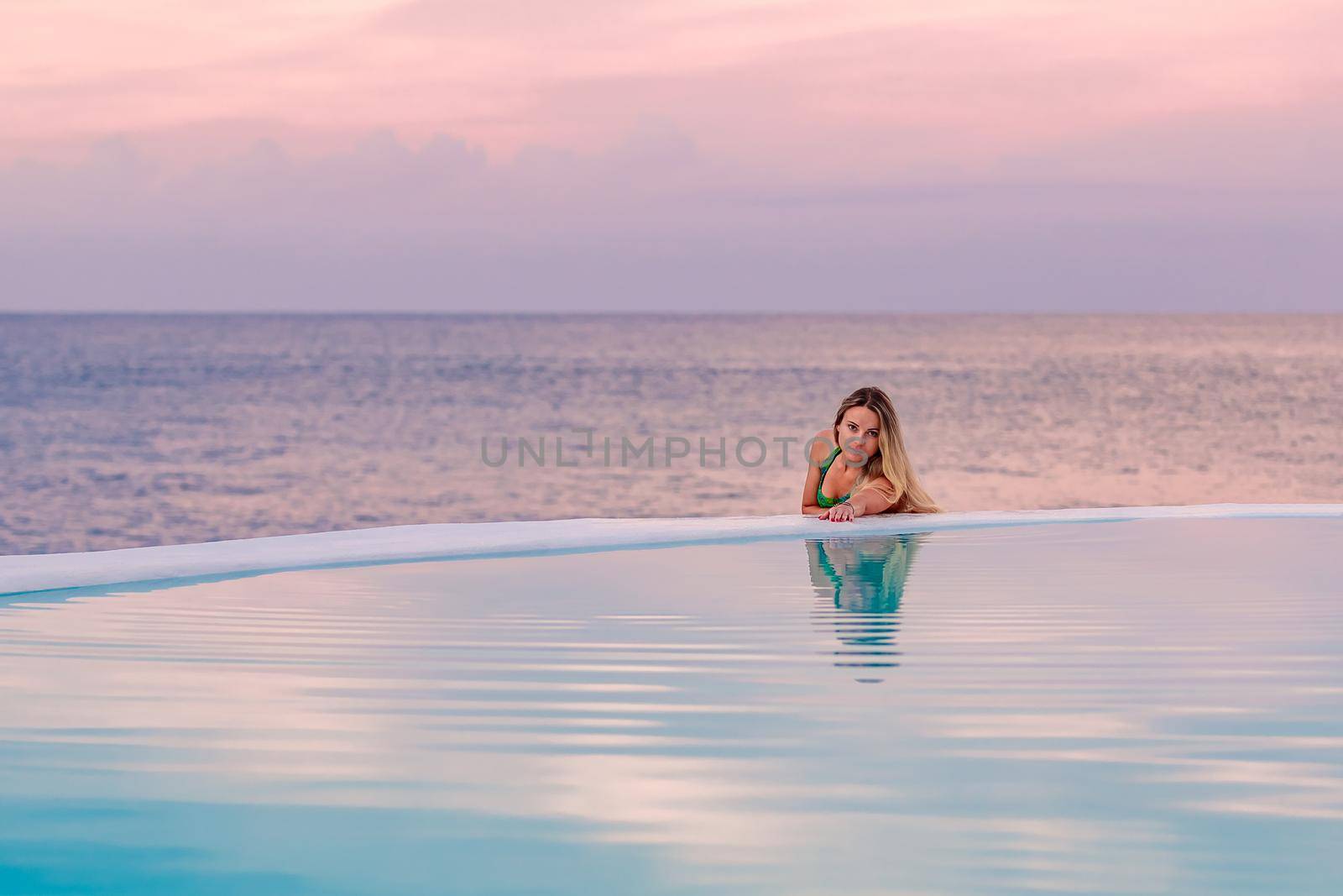 A young girl in a green swimsuit, at dawn by the sea pool, calmly and relaxed looking at her reflection in the water. Pink clouds in the background.
