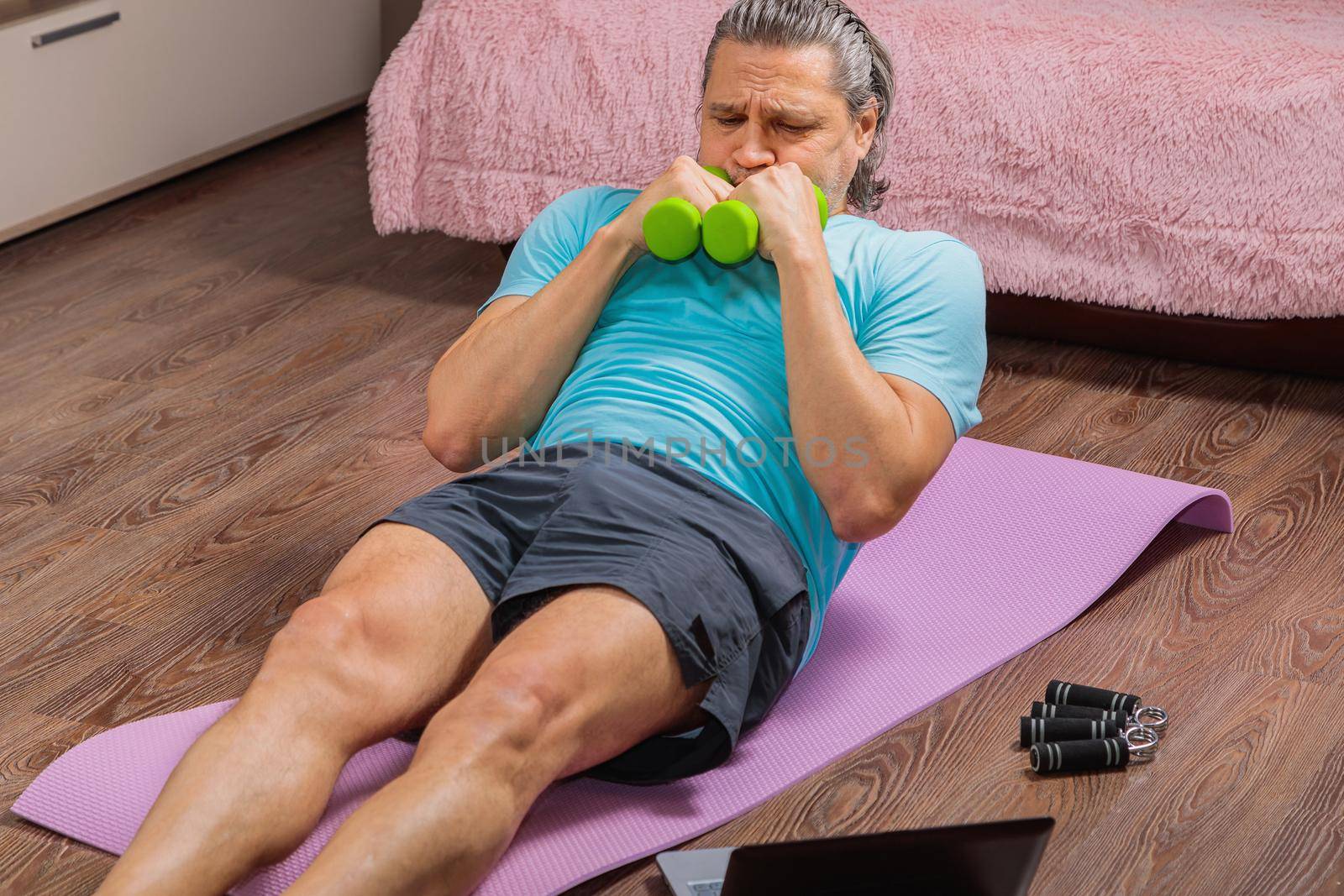 50-year-old man performs exercises while lying on the rug at home, looking at the computer. During a pandemic, a person trains in an apartment via the Internet.