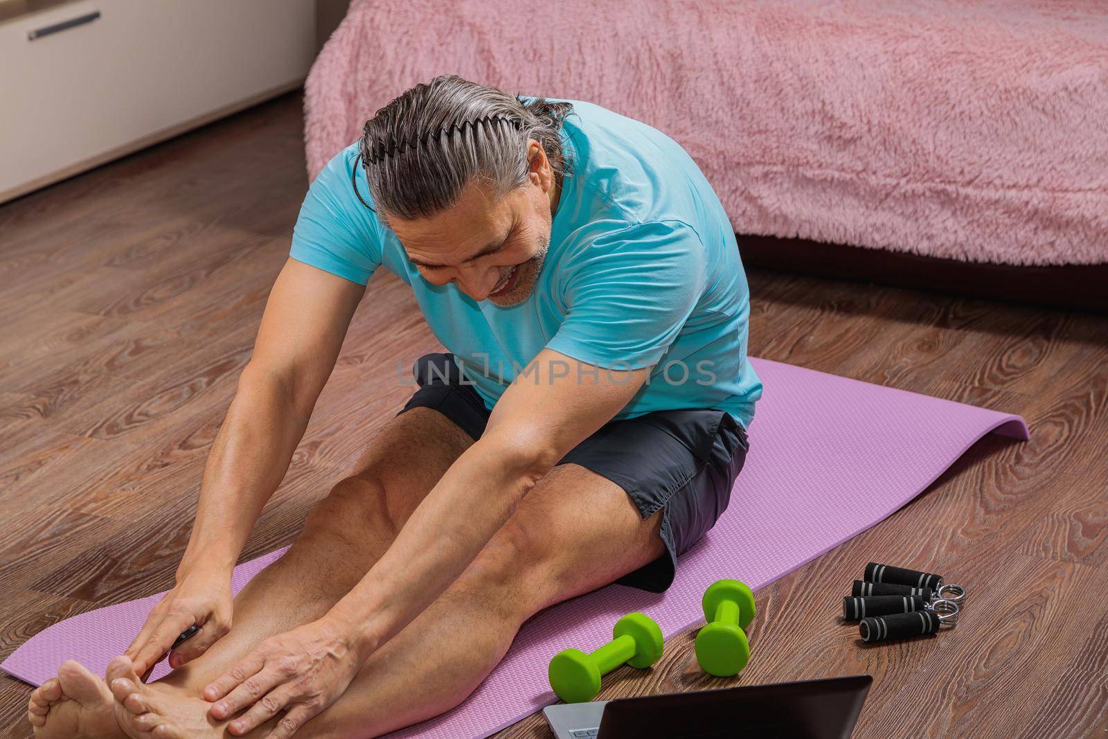 50-year-old man performs exercises while lying on the rug at home, looking at the computer. During a pandemic, a person trains in an apartment via the Internet.