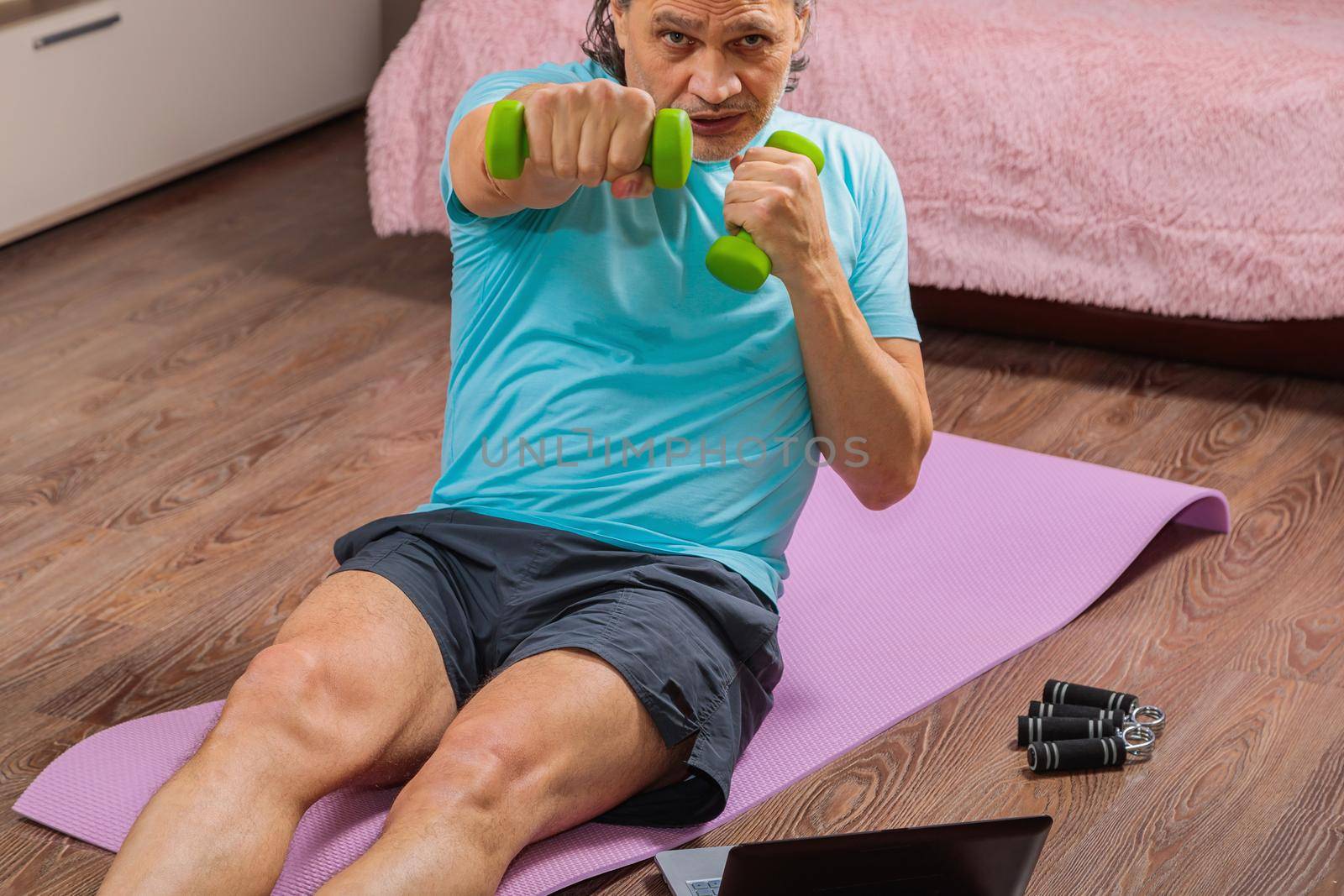 50-year-old man performs exercises while lying on the rug at home, looking at the computer. During a pandemic, a person trains in an apartment via the Internet.