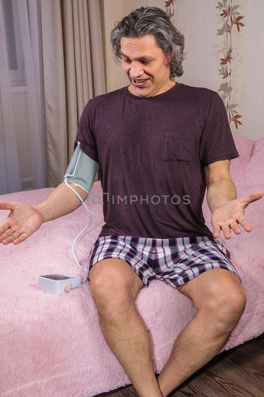 A 50-year-old man measures his blood pressure with a blood pressure monitor at home, sitting on his bed in his home clothes. Surprised by the instrument readings