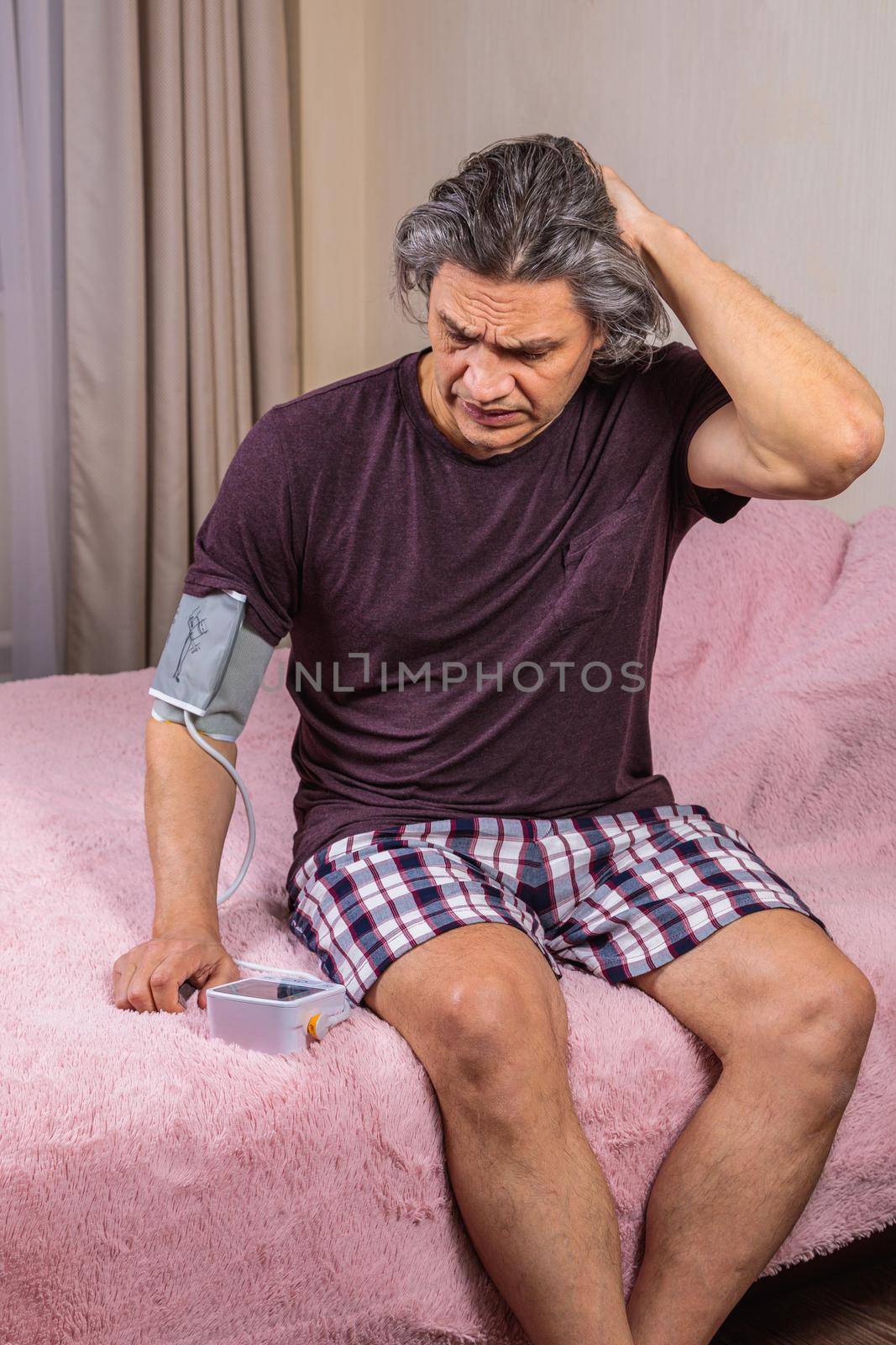 A 50-year-old man measures his blood pressure with a blood pressure monitor at home, sitting on his bed in his home clothes. Surprised by the instrument readings
