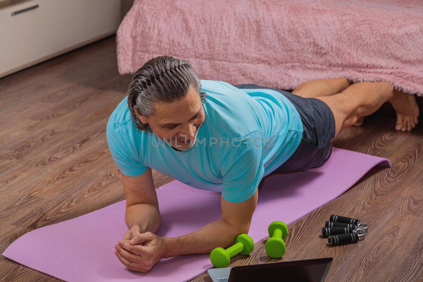 50-year-old man performs exercises while lying on the rug at home, looking at the computer. During a pandemic, a person trains in an apartment via the Internet.