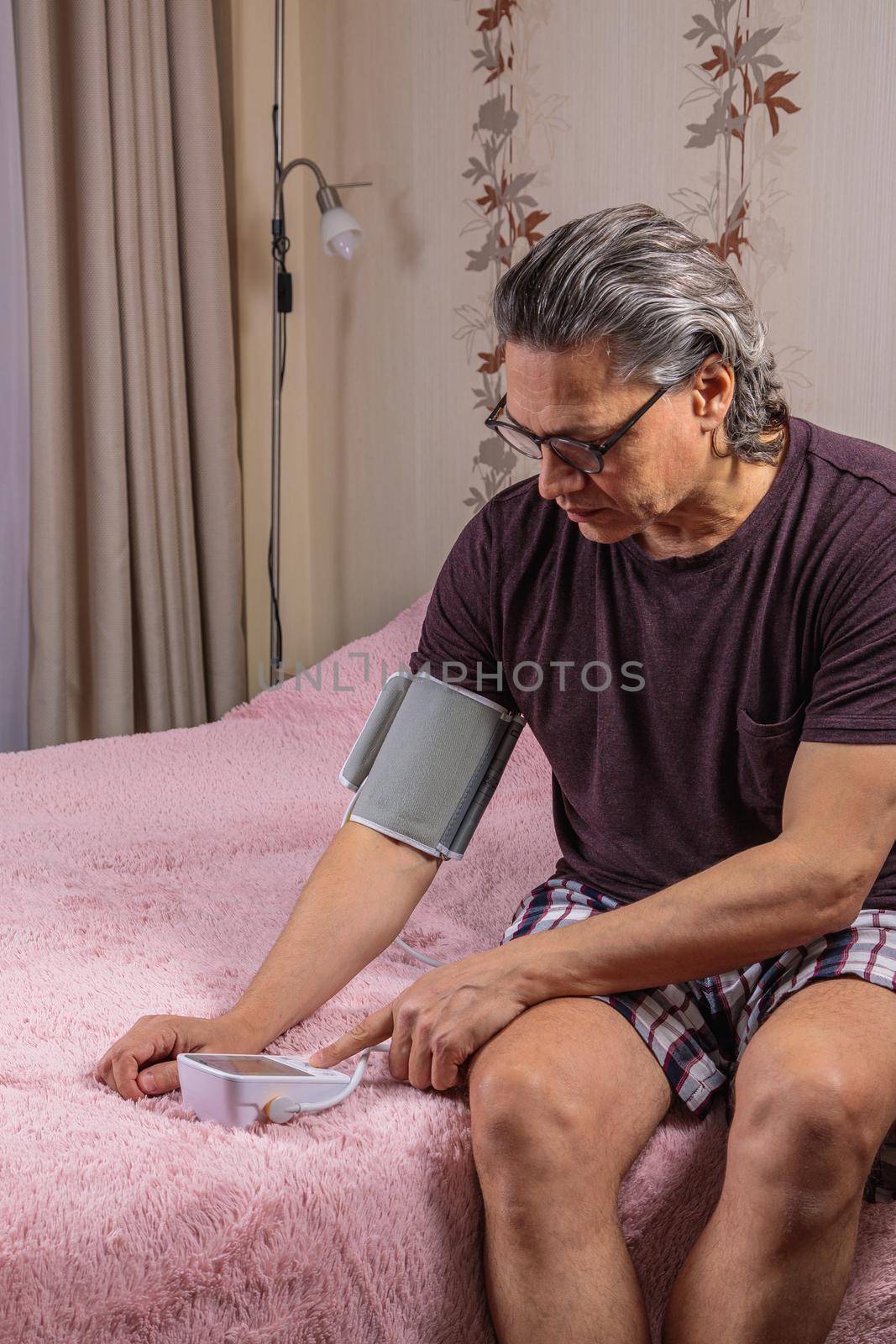 A 50-year-old man measures his blood pressure with a blood pressure monitor at home, sitting on his bed in his home clothes. Surprised by the instrument readings