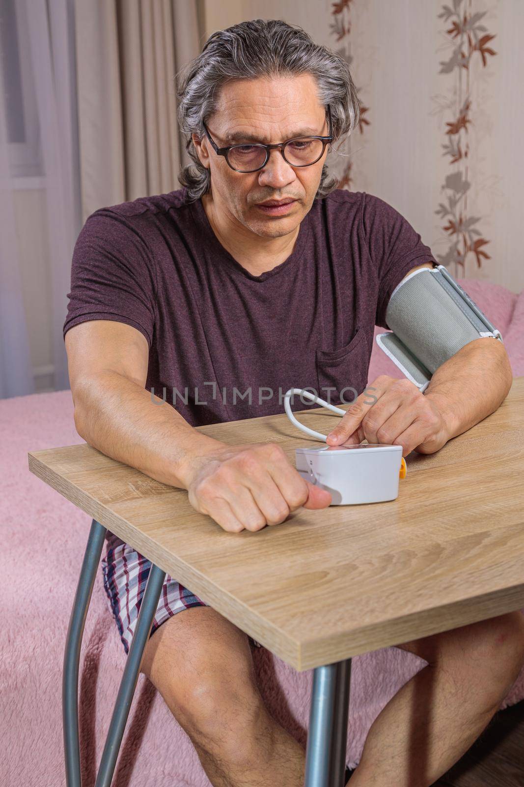 A 50-year-old man measures his blood pressure with a blood pressure monitor at home, sitting on his bed in his home clothes. Surprised by the instrument readings